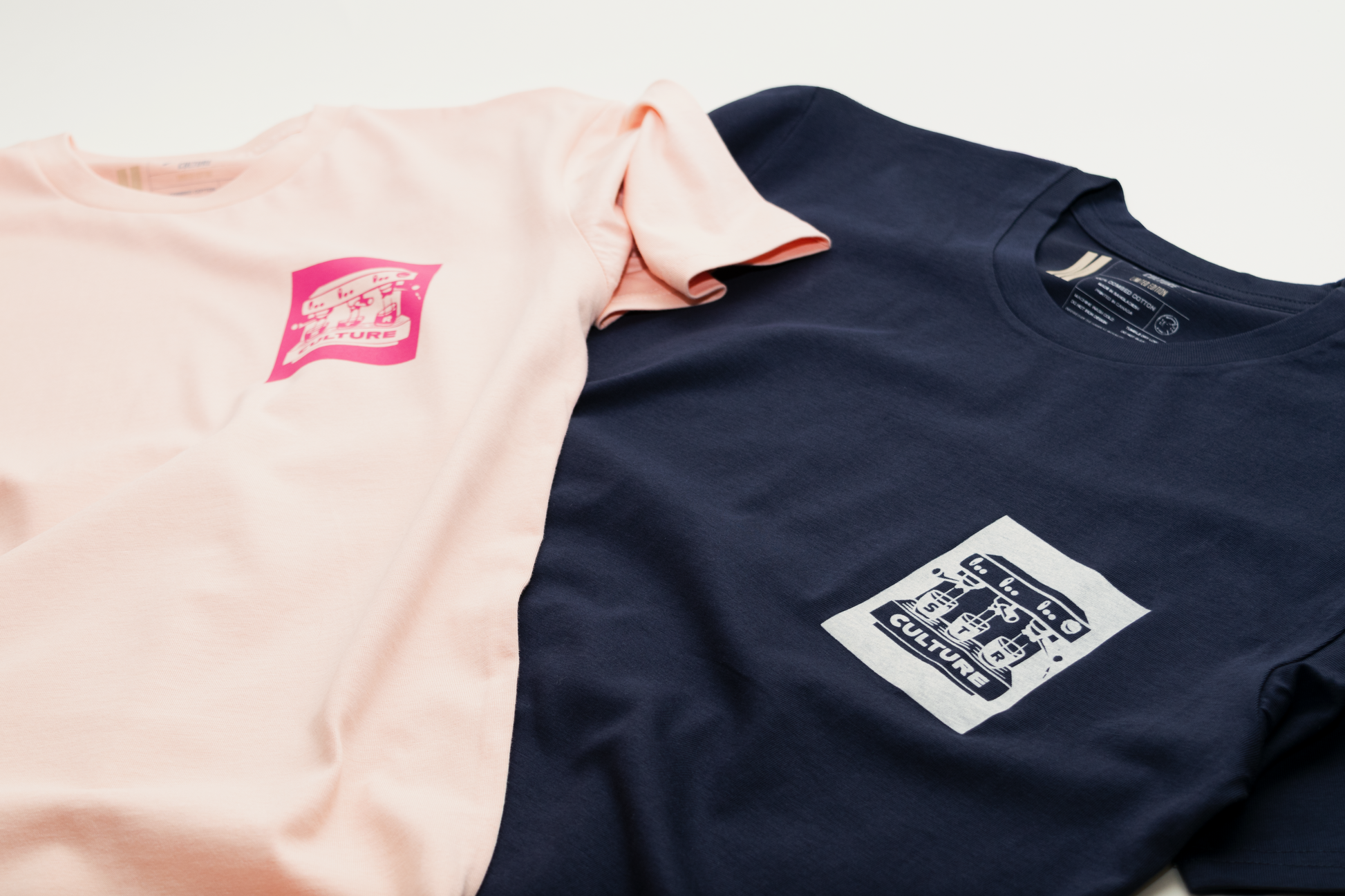 Two t-shirts, one pink and one navy, with logos on a light gray background